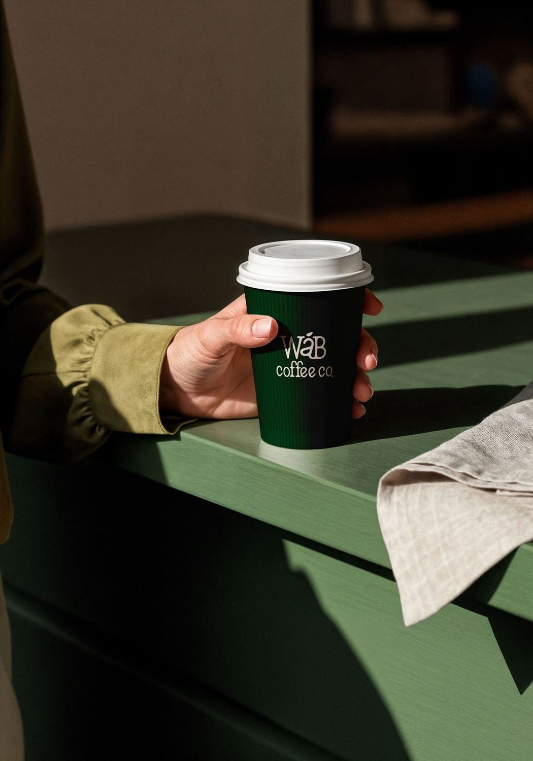 A hand holding a green WÁB branded coffee cup in sunlight